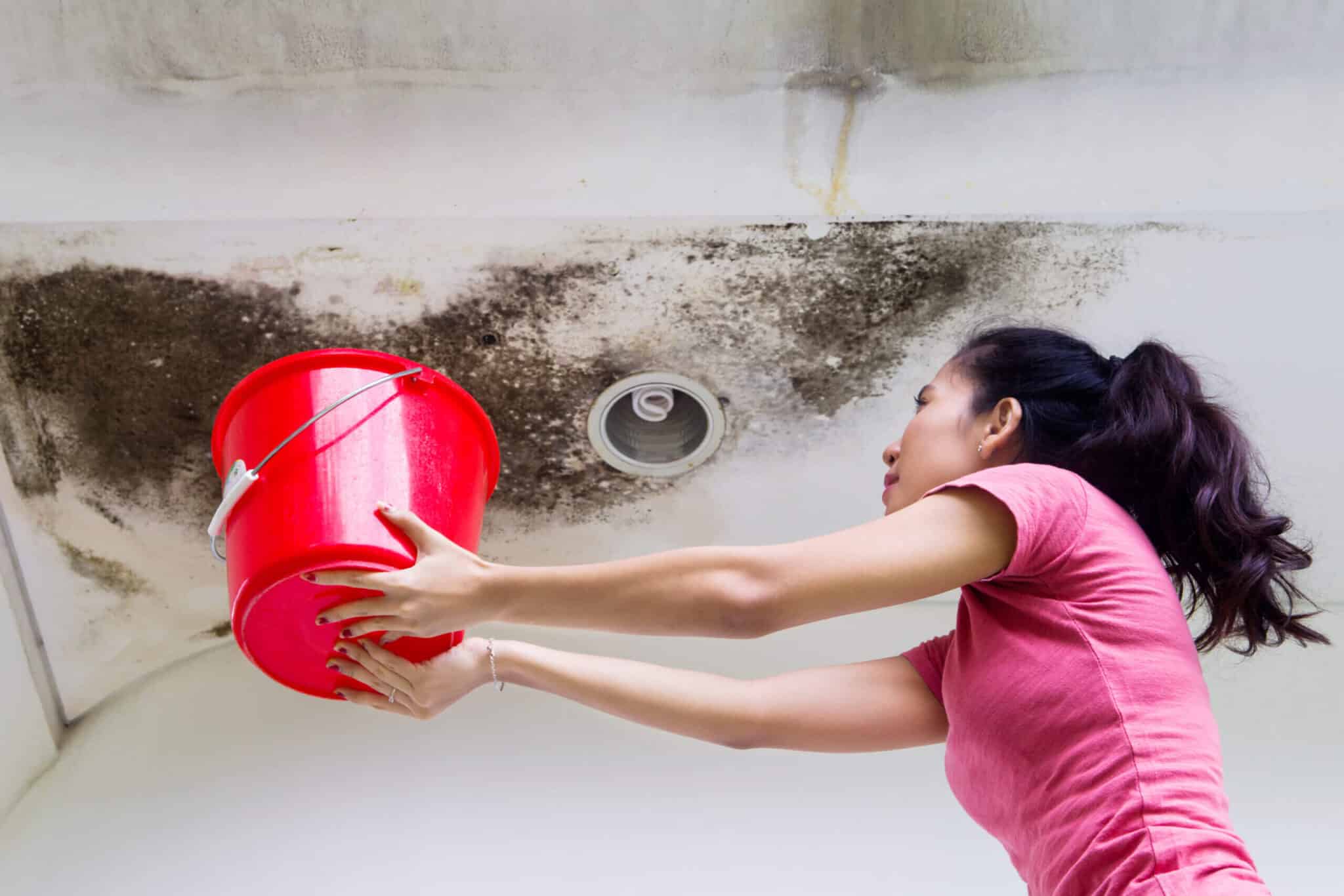 Photo of young woman holding a bucket while collecting drops rainwater from damaged ceiling